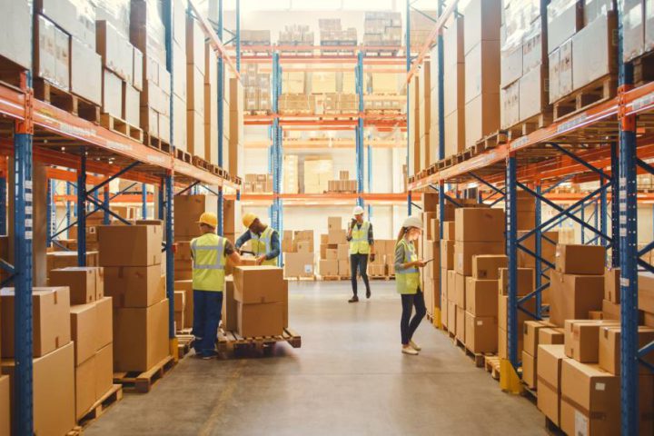 Four warehouse employees work inside a wide aisle. Two of them move boxes, while the others have a phone and clipboard.