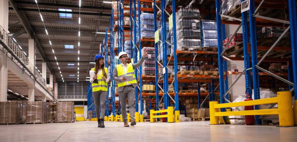 A man and woman, wearing safety vests and helmets while holding clipboards, walk around a warehouse.