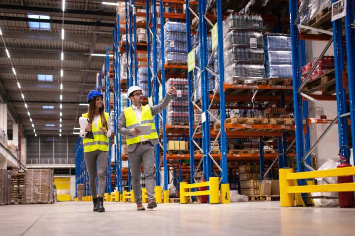 A man and woman, wearing safety vests and helmets while holding clipboards, walk around a warehouse.