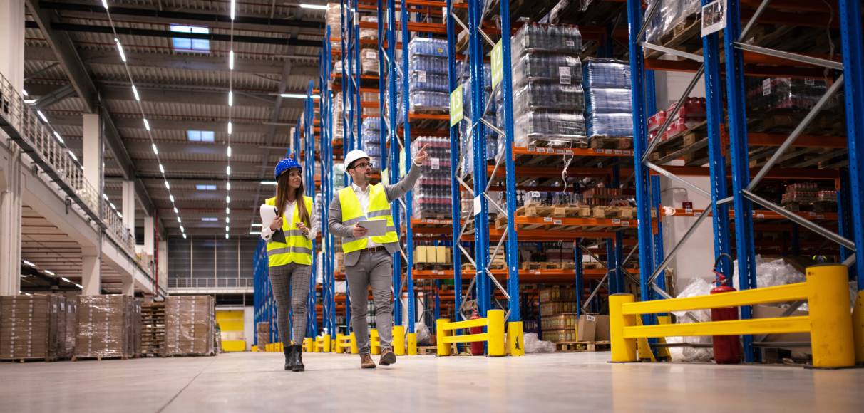 A man and woman, wearing safety vests and helmets while holding clipboards, walk around a warehouse.