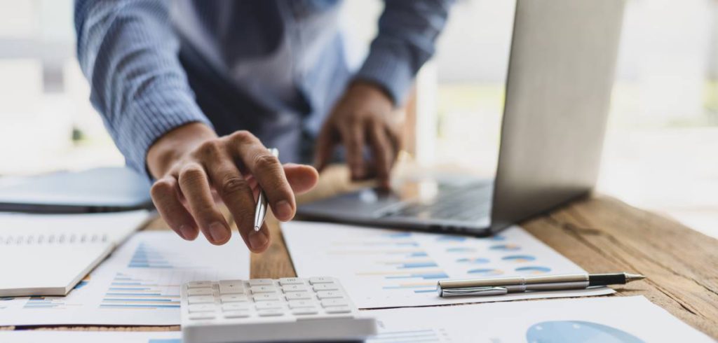 An executive working at his desk reaches over to his calculator. The desk also features papers with graphs and a laptop.