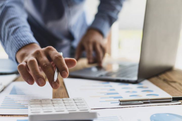 An executive working at his desk reaches over to his calculator. The desk also features papers with graphs and a laptop.