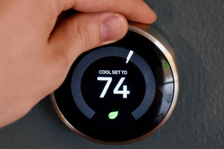 A hand turns the dial of a digital display of a thermostat reading "COOL SET TO 74" with a leaf symbol underneath.
