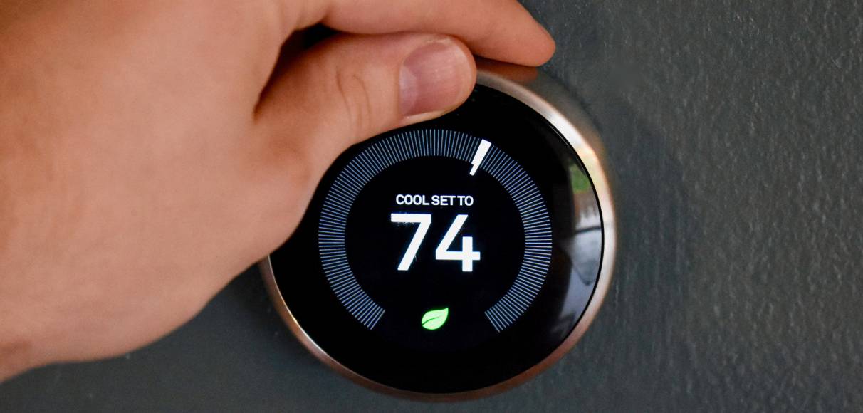 A hand turns the dial of a digital display of a thermostat reading "COOL SET TO 74" with a leaf symbol underneath.