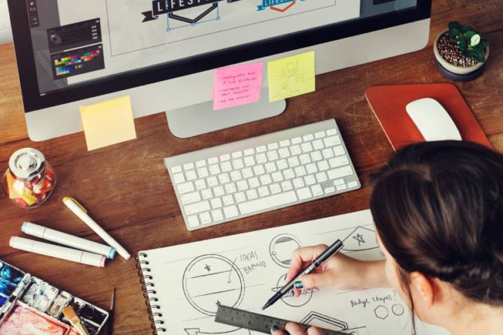 A designer is sitting at a computer with various images on the screen while drawing in a sketchpad.