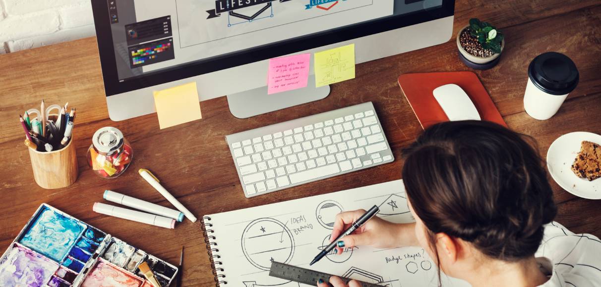 A designer is sitting at a computer with various images on the screen while drawing in a sketchpad.